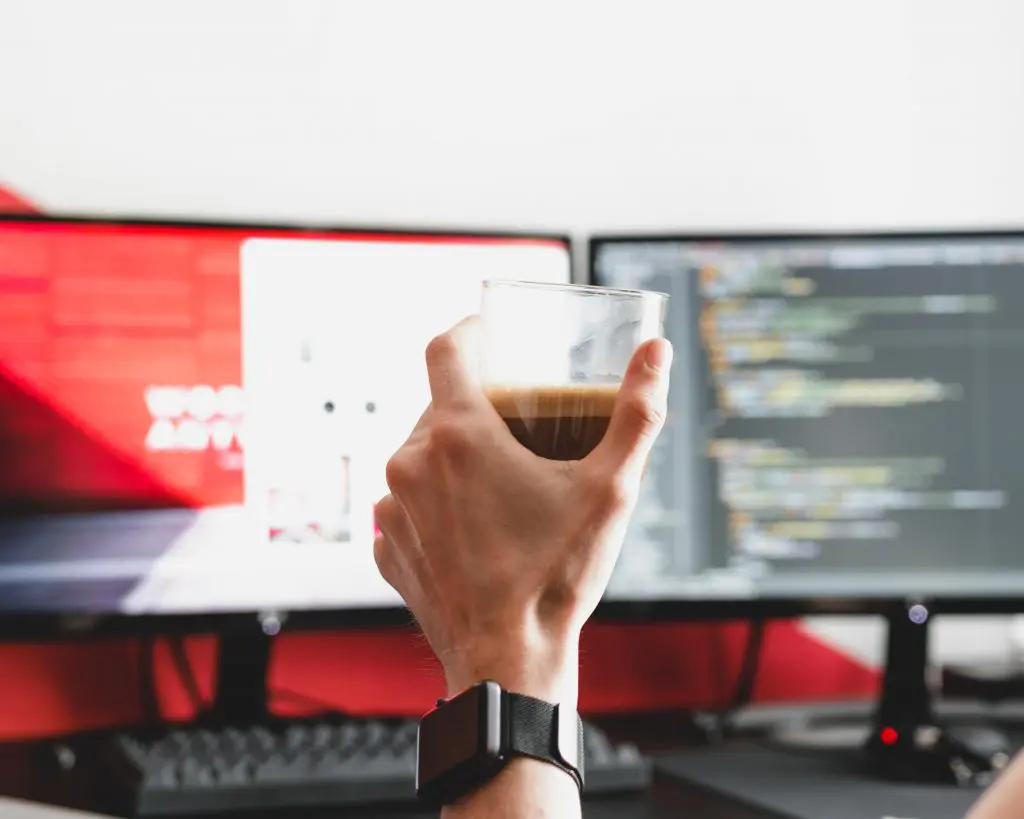 holding a cup of coffee in front of two screens showing code