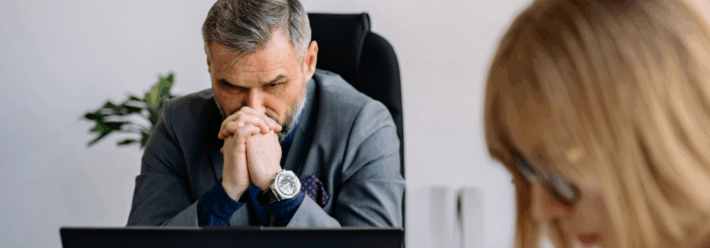 in besorgter Manager im Anzug sitzt mit gefalteten Händen am Schreibtisch und blickt ernst auf einen Bildschirm, während im unscharfen Vordergrund eine Kollegin arbeitet; Symbolbild für Stress und Herausforderungen bei Digitalisierungsprojekten.